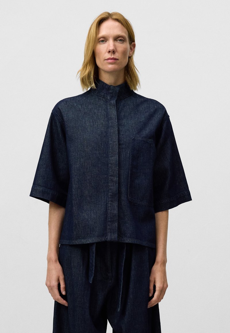 Camicia in denim scuro con colletto alto, maniche corte, una tasca a toppa frontale e vestibilità rilassata. Dettagli di cucitura e texture notevoli.
