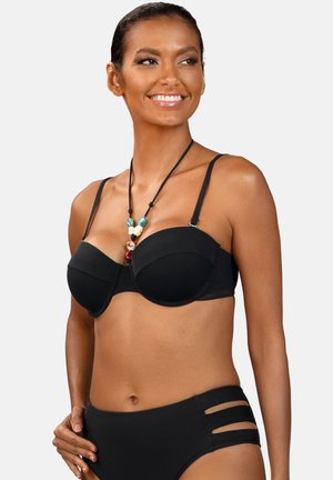 Uśmiechnięta kobieta w czarnym bikini z wycięciami na dole i naszyjnikiem z koralików, patrząca w bok na jednolitym tle.