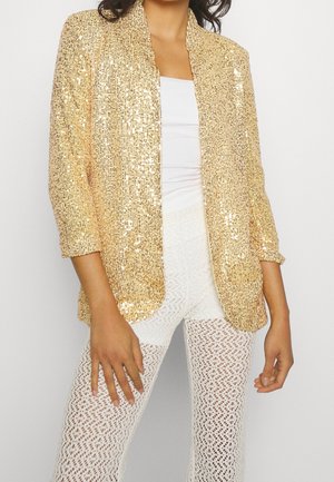 Frau trägt ein goldenes Pailletten-Blazer über einem weißen Oberteil und weißen gehäkelten Hosen, stehend vor einem einfachen hellen Hintergrund.