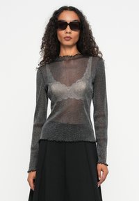 Must musta õhukese pika varrukaga pluus, millel on peen sära ja pitsäärised kaeluses ja hemis, kantud musta kihilise seeliku üle.