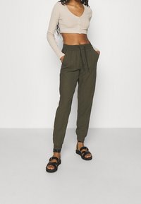 Hellbeige, taillierte Langarmbluse mit Vorderknöpfen, kombiniert mit olivgrünen Jogginghosen mit elastischem Bund und Bündchen. Schwarze Sandalen.