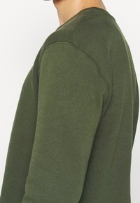 Sudadera verde oliva con cuello redondo, puños acanalados y costuras laterales. Confeccionada con una tela suave y texturizada. Detalle de costura visible.