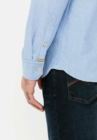 Lichtblauwe katoenen shirt met lange mouwen, knoopmanchetten en contrasterende gele stiksels. Gecombineerd met donkere jeans die de detail van de zijzakken laten zien.
