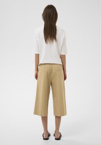 Femme portant un haut blanc à manches courtes, un pantalon large beige court et des sandales noires à enfiler, debout devant un fond clair uni.