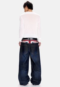 Άτομο με κοντά σκούρα μαλλιά που φοράει διάφανο λευκό πουκάμισο με μακριά μανίκια και φαρδιά τζιν παντελόνια με ζώνη στην μέση με το σχέδιο της σημαίας του Ηνωμένου Βασιλείου.