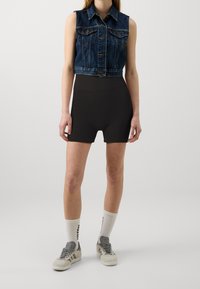 Denim mouwloos jasje met knopen aan de voorkant, zwarte geribbelde high-waisted shorts en grijze sneakers met witte sokken met tekst.