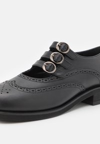 Chaussure brogue en cuir noir avec trois brides à boucle argentée et motifs décoratifs perforés sur le dessus et l’orteil.