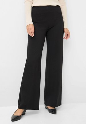 Schwarze weit geschnittene Hose aus glattem Stoff, mit hoher Taille, einem leichten Schlitz am Saum, kombiniert mit schwarzen flachen Schuhen.