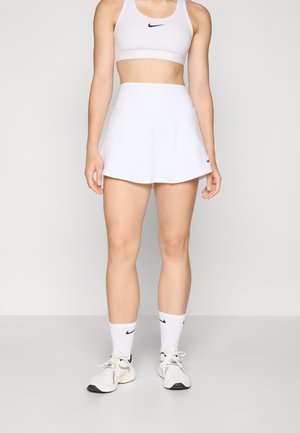 Žena nosící bílý sportovní top Nike, bílou tenisovou sukni, bílé ponožky Nike a bílé sportovní boty stojící na šedé podlaze.