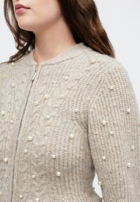 Mulher vestindo um suéter de malha bege decorado com pérolas pequenas e grandes espalhadas, apresentando um fecho de zíper na frente.