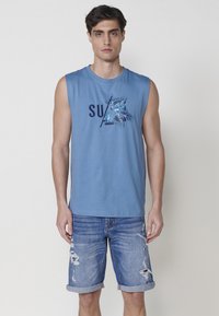 Svetlo modra brezrokavna majica s potiskom, ki prikazuje "SURF" in palmove liste, v kombinaciji z raztrganimi jeans kratkimi hlačami, zvitimi na robu.