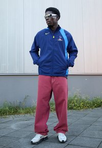 Marineblå og lyseblå vindjakke med logo, røde løse bukser og hvide sneakers med sorte detaljer. Model iført briller står mod en væg.