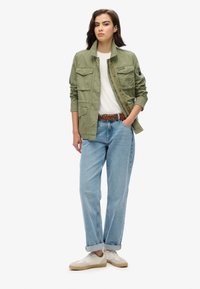 Groene katoenjack met meerdere zakken, witte t-shirt, lichtblauwe spijkerbroek en beige sneakers, met opgerolde mouwen en een gevlochten riem.