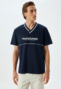 Marineblå t-shirt med v-hals og hvidt læg, med teksten "COLLEGE LEAGUE EST. 1988" trykt i midten. Korte ærmer og blødt bomuldsstof.