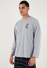 Cască gri cu mânecă lungă, realizată dintr-un material moale, având un imprimeu colorat cu desene animate pe pieptul stâng. Modelul poartă pantaloni de pijama cu carouri.
