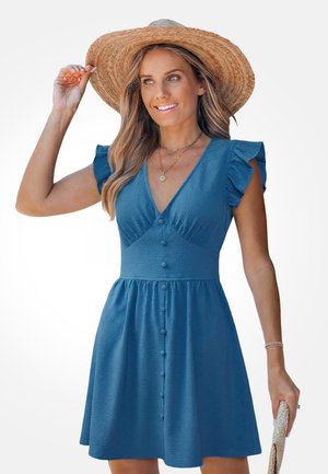 Kobieta ubrana w niebieską sukienkę z guzikami z falbankami na rękawach i szerokim słomkowym kapeluszem, trzymająca kapelusz jedną ręką.