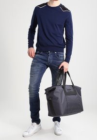 Borsa a duffel grigia realizzata in tessuto testurizzato, con accenti neri e una cerniera superiore. Indossata con un maglione blu navy e jeans blu. Sneakers bianche visibili.