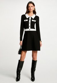 Vestido de punto en blanco y negro con cuello tipo blazer, botones al frente, dos bolsillos en el pecho y falda acampanada. Llevado con botas negras hasta la rodilla.