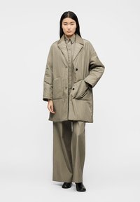 Γυναίκα που φοράει φαρδιά μπεζ κοστούμι με υπερμεγέθη σακάκι και παντελόνια με φαρδιά μπατζάκια, στέκεται μπροστά σε απλό ανοιχτό φόντο.