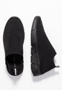 Baskets en maille noires à enfiler avec semelles texturées et bordures élastiques autour des chevilles, montrées vues de dessus et de côté sur fond blanc.