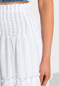 Saia branca com padrões intrincados em azul claro, apresentando uma cintura franzida e barra em babados. Tecido suave e texturizado.