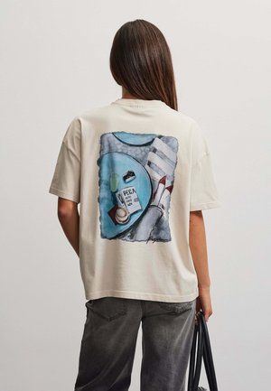 Persona con cabello largo y castaño que lleva una camiseta beige con una ilustración de pies, mesa con café, cámara y nota vista desde arriba, sosteniendo una bolsa negra.