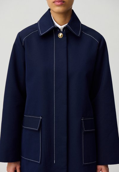 Manteau bleu marine avec un col haut, fermeture à boutons dorés et couture blanche. Présente deux poches avant avec rabats. Texture de tissu lisse.