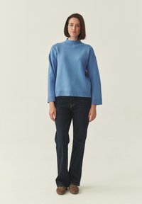 Pull oversize bleu clair avec un col montant, des poignets et un ourlet côtelés. Associé à un jean évasé en denim foncé et des chaussures marron.