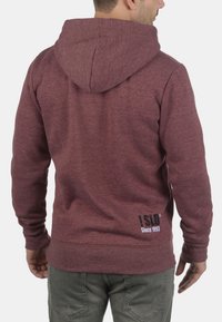 Bordeaux sweatshirt med hætte, fremstillet af et tekstureret stof, med ribbede cuffs og et logo med teksten "Siden 1993" printet i hvidt på den nederste del af ryggen.