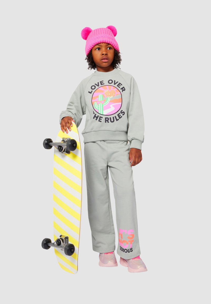 Enfant en tenue grise avec un chapeau rose tenant un skateboard à rayures jaunes, se tenant devant un fond gris clair.