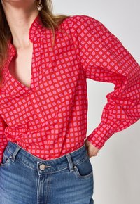 Blusa rossa a fantasia con design floreale rosa, colletto a scollo a V e maniche a palloncino, abbinata a jeans blu.