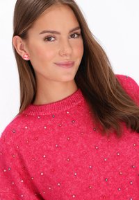 Pullover rosa brillante con una finitura testurizzata, adornato con perline multicolori. Accompagnato da orecchini a righe, che presentano un design circolare.