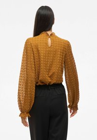 Blusa color senape con tessuto testurizzato e stampato, maniche lunghe con polsini a volant e un'apertura a occhiello sul retro.