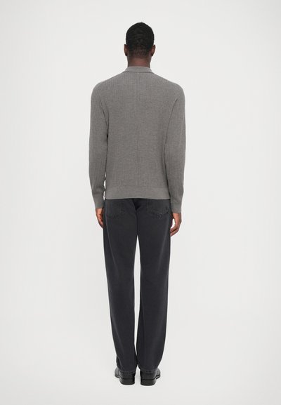 Grijs geribbeld gebreid sweater met lange mouwen, met een ronde hals. Draagt bij donkere broek en zwarte schoenen. Effen achtergrond.