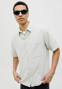 Camisa de manga corta de color gris claro con tejido texturizado, botonadura frontal, bolsillo en el pecho y botones azules. El modelo lleva gafas de sol y un collar de cadena.