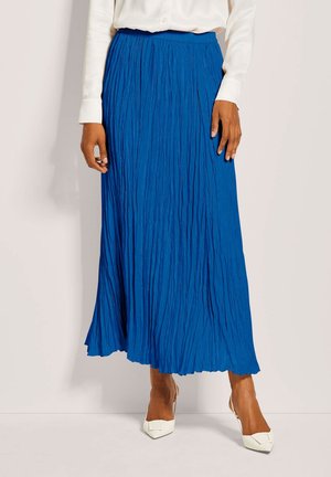 Vrouw in een witte blouse, heldere blauwe geplooide midi rok en witte puntige slingback hakken op een effen achtergrond.