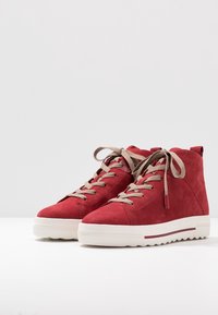 Czerwona zamszowa wysokoprofilowa sportowa obuwie z beżowymi sznurowadłami, białą teksturowaną gumową podeszwą i lekkim obcasem. Widoczne detale bocznego przeszycia.