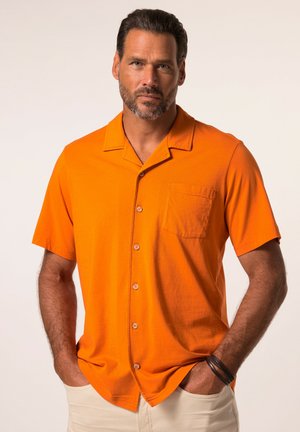 Man met baard die een feloranje kortemouwen overhemd met knopen draagt en lichte broek, handen in de zakken.