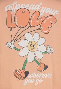 Perzikkleurig T-shirt met een cartoonachtige bloemenkarakter dat hartvormige ballonnen vasthoudt, met de tekst "Verspreid je LIEFDE waar je ook gaat".