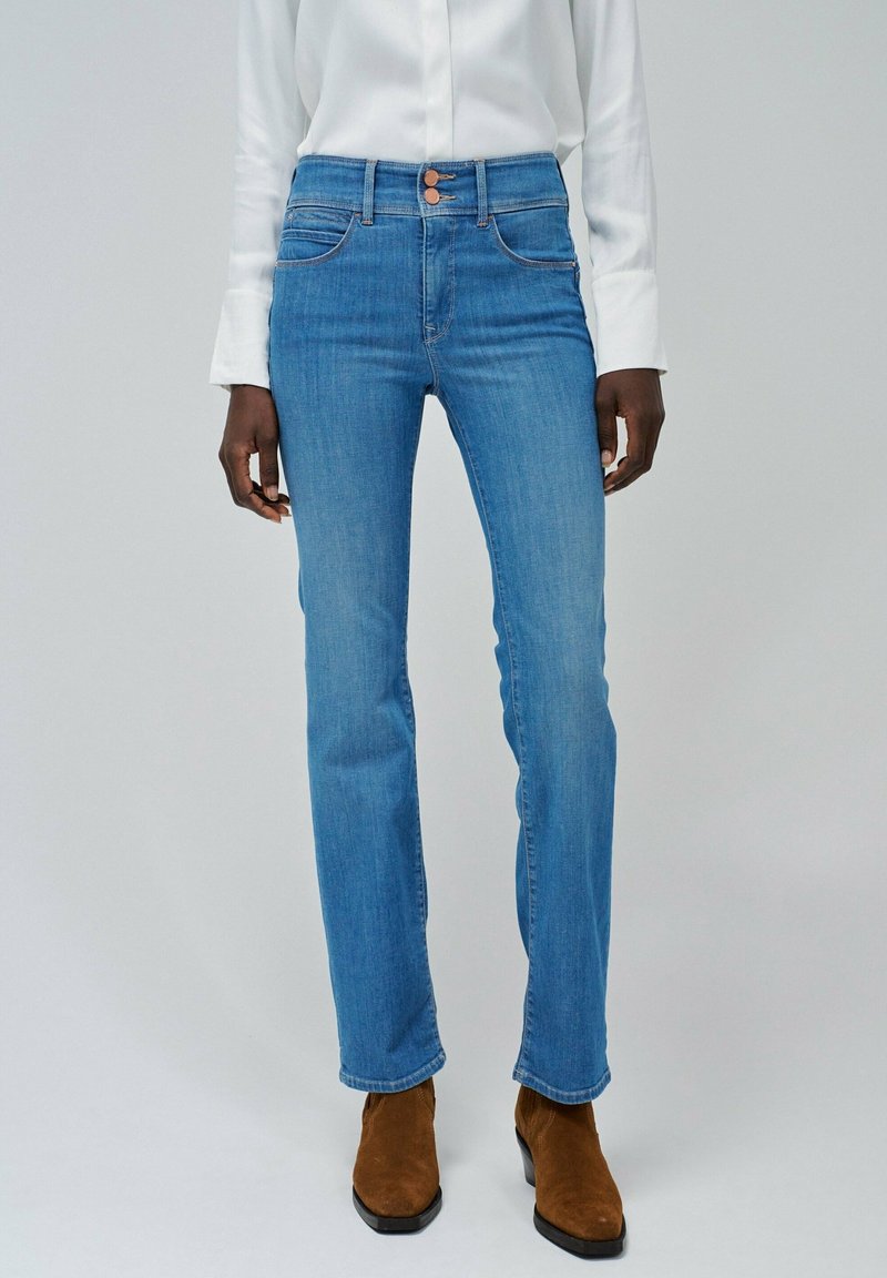 Personne portant un jean bleu clair taille haute avec deux boutons, associé à un haut blanc et des bottines en daim marron, debout devant un fond uni.