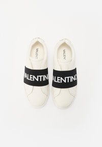 Sneaker bianco con un elastico nero che presenta "VALENTINO" in testo bianco. Punta rotonda e texture in pelle liscia. Design minimalista.