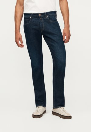 Mand iført mørkeblå jeans med lige ben og hvide sneakers, stående mod en ensartet lys baggrund.