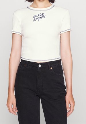 Woman wearing a white cropped short-sleeve top with "guess originals" text and high-waisted black jeans, standing against a plain background.