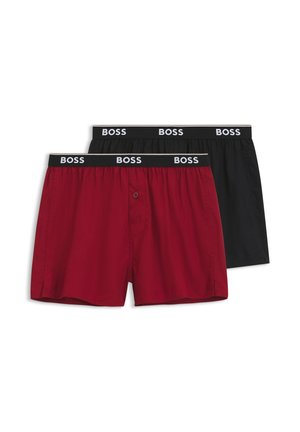Uomo muscoloso che indossa boxer rossi BOSS con elastico nero con marchio, in piedi davanti a uno sfondo bianco uniforme.