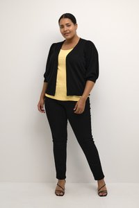Cardigan noir à manches bouffantes, porté sur un top jaune. Un pantalon noir slim et des sandales noires à lanières complètent la tenue.