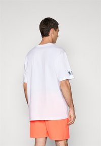 Mann som har på seg en enkel hvit oversized t-skjorte og knalloransje shorts, med ryggen mot en lys grå bakgrunn.