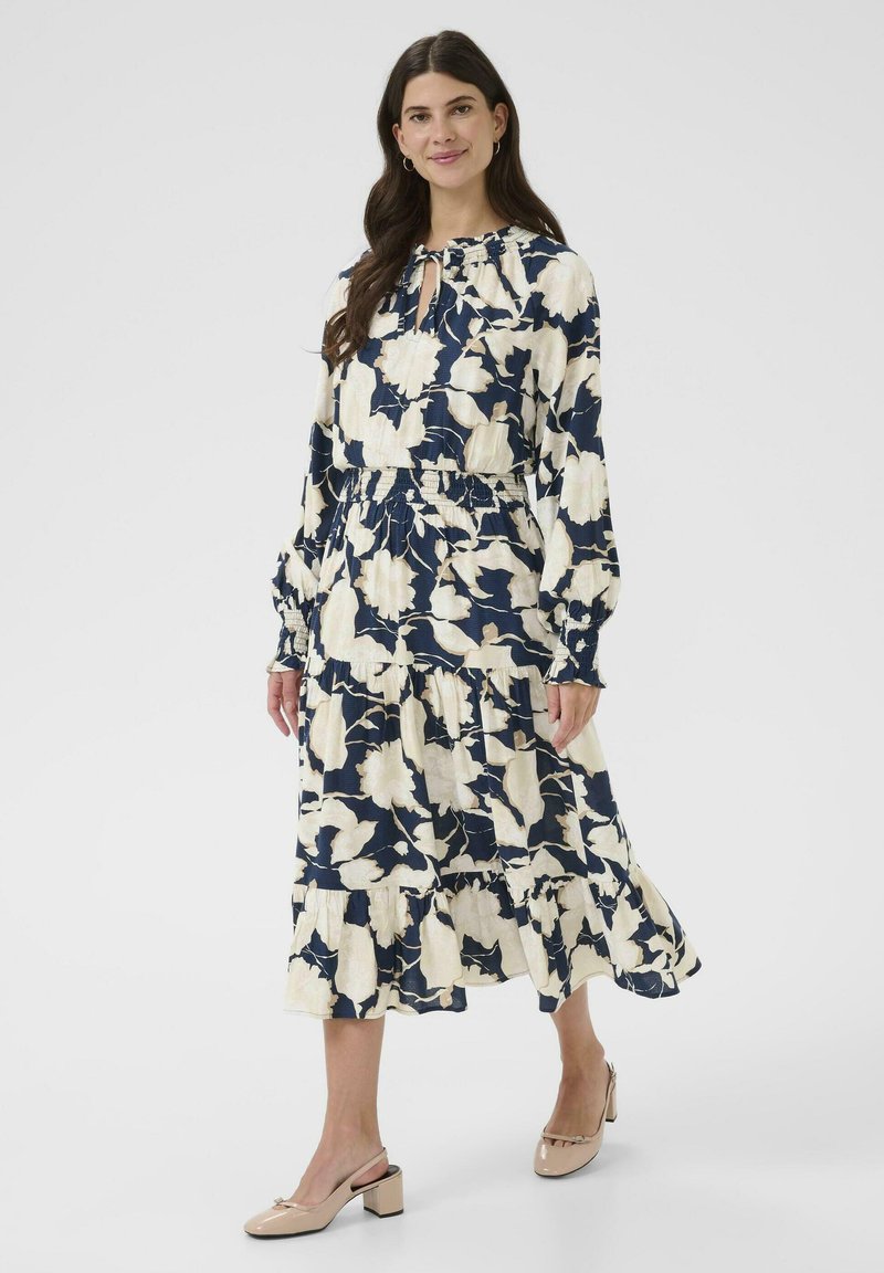 Bloemenmaxi-jurk in marineblauw en crème, met een verzamelde taille, lange mouwen, een sleutelgatdecolleté en een gelaagde rok. Gecombineerd met beige block-heeled schoenen.