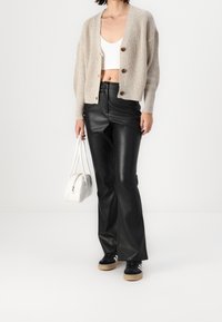 Beige stickad cardigan med tre knappar, creamfärgad kort topp, svarta läderbyxor med vida ben, vit handväska och svarta sneakers med gummisula.