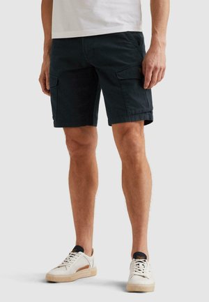 Mann trägt schwarze Cargo-Shorts und weiße Turnschuhe, steht vor einem einfarbigen Hintergrund.