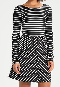 Robe à manches longues en rayures noires et blanches. Corsage ajusté avec une jupe évasée. Rayures horizontales en haut, passant à des rayures diagonales à la taille.